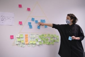 Person pointing to post-it note in centre of cluster, identifying common theme as part of design workshop.
