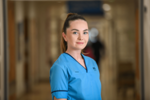 Image of Louise wearing blue NHS uniform in hospital corridor. 