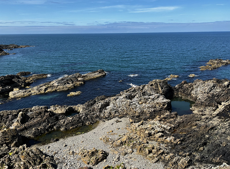 Seascape with clear blue skies and seas, rocky coastline in the foreground.